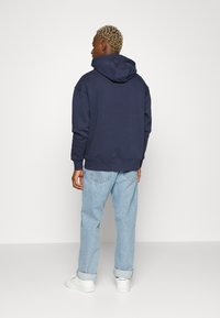 Sudadera azul marina con capucha ajustable, corte holgado, puños y dobladillo acanalados, combinada con jeans de un azul claro y zapatillas blancas.