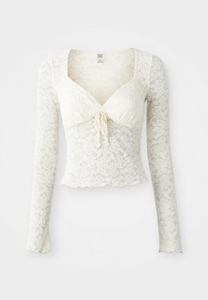 Crop top en dentelle crème à manches longues, avec un décolleté en V, un détail à nouer à l'avant, un corps ajusté et un ourlet échancré.
