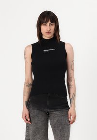 Juodas be rankovių golfo megztinis su balta siuvinėta "KARL LAGERFELD JEANS" logotipu kairėje pusėje, derinamas su tamsiai pilkomis džinsomis.