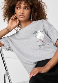 Grå bomulds t-shirt med oprullede ærmer, der har et trykt grafisk motiv af en figur i en hvid kjole og pink støvler. Der er detaljer som stjerner og tekst.