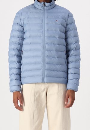 Homme portant une veste matelassée bleu clair zippée, associée à un pantalon beige clair, debout devant un fond uni.