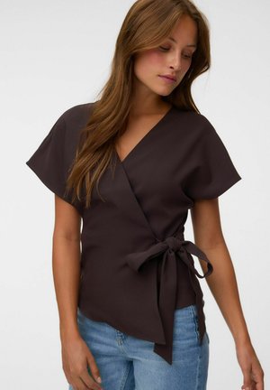 Femme aux longs cheveux bruns portant un chemisier cache-cœur foncé à manches courtes noué à la taille et un jean bleu clair.