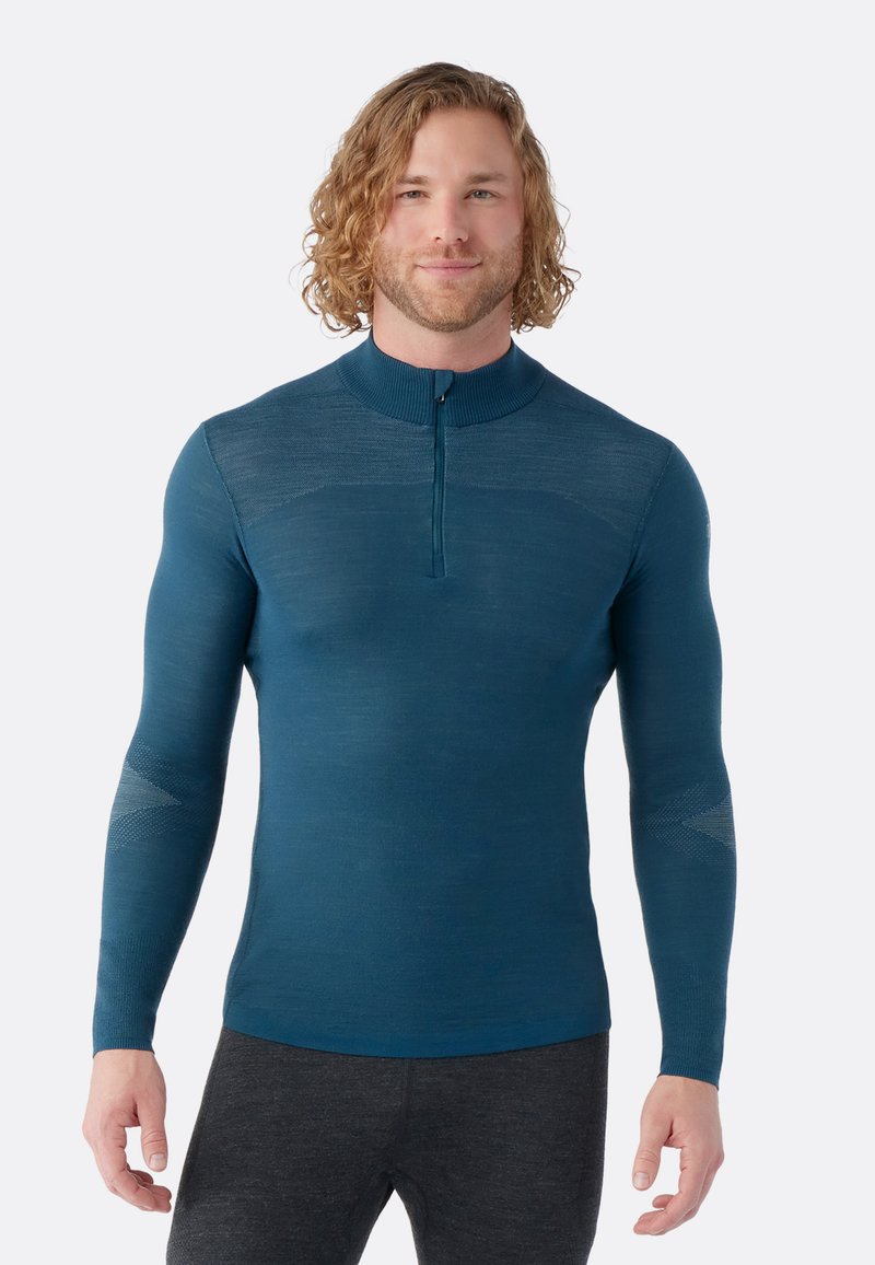 Teal sportovní dlouhý rukáv s vysokým límcem a polovičním zipem; má strukturované panely na rukávech a těle pro zvýšenou flexibilitu.