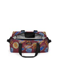 Cestovní taška z vínově červené látky s různými barevnými nášivkami a logy. Viditelné černé popruhy a vnitřní úložná kapsa.