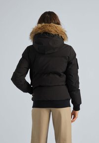 Sort pufferjakke med aftagelig pelsforet hætte, polstret design, strikkede manchetter og sidelommer. Båret over beige bukser.