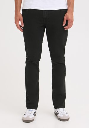 Homme portant un jean slim noir et des baskets blanches avec des bandes noires, debout devant un fond blanc uni.