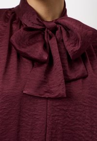 Bordeaux blouse met een grote strik bij de kraag, gemaakt van textuurstof met een subtiele glans, valt zachtjes rond de halslijn.
