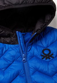 Modrá prošívaná bunda s černou kapucí, se zapínáním na přední straně a černým logem. Textura je hladká a polstrovaná.