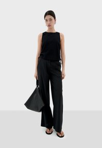 Schwarzes, strukturiertes, ärmelloses Oberteil mit Kordelzug an den Seiten, kombiniert mit schwarzen, gestreiften, weiten Hosen. Das Model hält eine große schwarze Tote-Tasche.