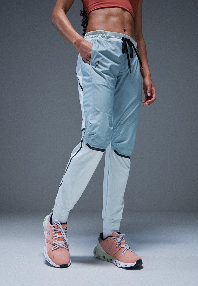 Hellblaue Sporthosen mit schwarzen Akzenten, elastischem Bund und schmal zulaufenden Beinen, kombiniert mit pink- und graufarbenen Turnschuhen.