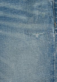 Tessuto in denim blu chiaro con una piccola toppa rattoppata e cucita vicino a una cucitura verticale sul lato destro.