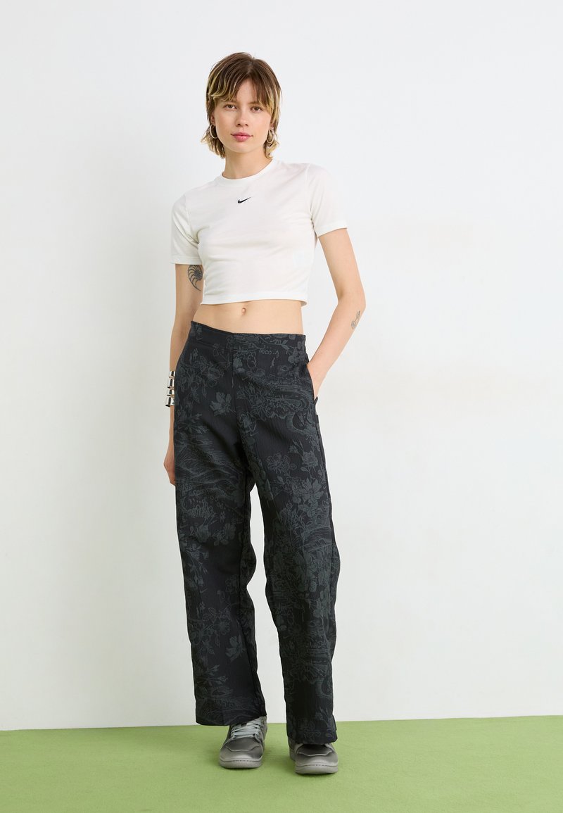 Pantalones de pierna ancha con estampado floral negro, fabricados con un material ligero. Combinados con una camiseta corta blanca de Nike y unas zapatillas grises.