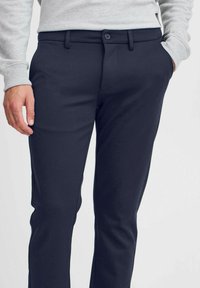 Pantalones azul marino fabricados con tejido suave, con cierre de botón, dos bolsillos frontales y un corte entallado. Textura sutil y diseño aerodinámico.