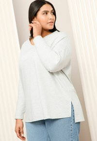 Ljusgrå långärmad topp med V-ringning, med sidofickor. Matchad med blå denimjeans som visar en avslappnad passform och mjuk textur.