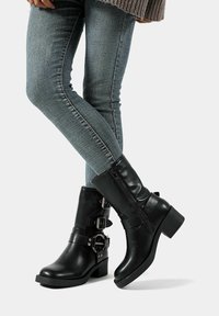 Stivali neri in pelle fino alla caviglia con tacco chunky, due dettagli con fibbie e una cerniera laterale, abbinati a jeans slim azzurri.