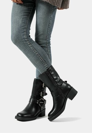 Stivali neri in pelle fino alla caviglia con tacco chunky, due dettagli con fibbie e una cerniera laterale, abbinati a jeans slim azzurri.