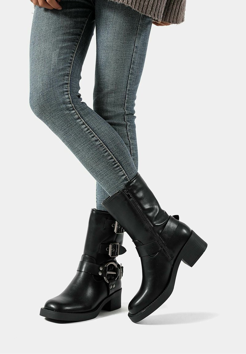 Stivali neri in pelle fino alla caviglia con tacco chunky, due dettagli con fibbie e una cerniera laterale, abbinati a jeans slim azzurri.