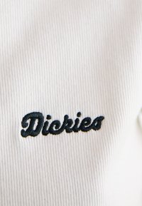 Tessuto bianco a righe con logo "Dickies" ricamato in nero. La trama è morbida con dettagli a righe ben visibili.