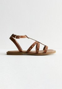 Brun konstläder sandal med platt sula, justerbart ankelsnöre och flera korsade remmar med guldnitar. Slät yta, minimalistisk design.