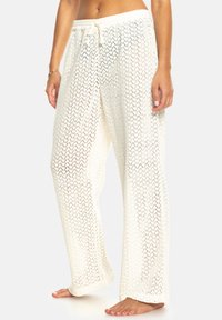 Witte, lichte, luchtige gebreide broek met een losse pasvorm, voorzien van een trekkoord in de taille en een ingewikkeld ruitpatroon over de hele broek. Voeten zijn bloot.