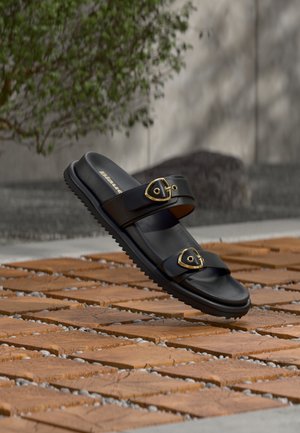 Črna sandal z dvema zlatima zaponkama, ki lebdi nad lesenim tlemi iz ploščic, v ozadju zamegljeno zeleno grmičevje in sivi zid.