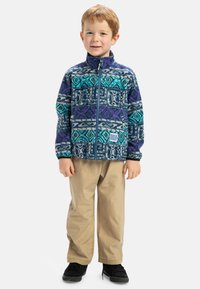 Veste en polaire avec des motifs géométriques violets et bleus canard, fermeture éclair complète, col montant ; associée à un pantalon beige et des baskets noires.