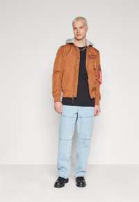 Jachetă bomber maro cu glugă gri, cu fermoar în față și detalii cu patch-uri. Purtați peste un tricou negru, asortată cu pantaloni din denim bleu deschis și pantofi negri.