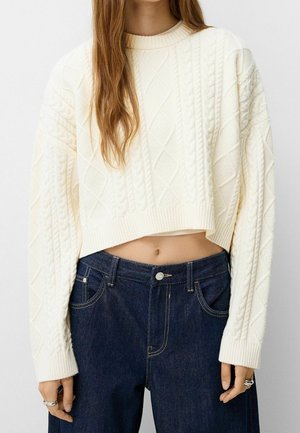 Femme portant un pull court blanc torsadé à manches longues et un jean foncé taille haute, montrant un ventre nu.