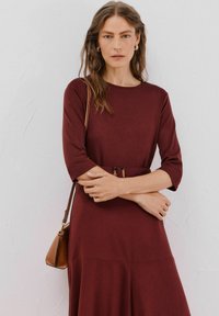 Burgunderfarbenes Langarmkleid mit Rundhalsausschnitt, tailliertem Gürtel und fließendem Rock. Accessorized mit einer kleinen braunen Handtasche.