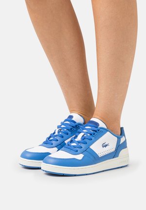 Baskets basses bleu et blanc à lacets portées aux pieds, avec un logo sur le côté et des semelles blanches sur un fond uni.