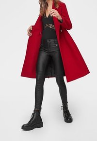 Röd ullkappa, svart spetsbodysuit, svarta läderbyxor och svarta snörboots. Kappan är lång och åtsittande, med knappar som detalj.