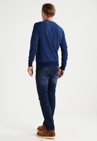 Námorně modrý svetr s dlouhými rukávy a žebrovaným lemem, spárovaný s tmavými džíny. Model otočen zády, předvádí uvolněný střih a hladkou strukturu.