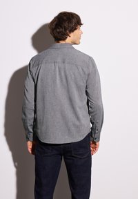 Camicia button-down grigia a maniche lunghe con finitura testurizzata, caratterizzata da un orlo arrotondato, dettagli delle cuciture e polsini con bottoni, abbinata a jeans scuri.