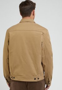 Beige Jacke aus glattem Stoff, mit einem Kragen, sichtbaren Nähten, Knopfakzenten und geradem Schnitt.