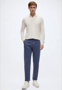 Φωτεινή μπεζ μακρυμάνικη μπλούζα πόλο, μπλε σκούρο παντελόνι και λευκά αθλητικά παπούτσια. Απλός σχεδιασμός με άνετη εφαρμογή και διακριτικές λεπτομέρειες στον γιακά.