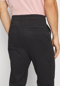 Svarta byxor med en slät yta, med två bakfickor och rak passform, kombinerade med en ljusrosa skjorta i midjan.