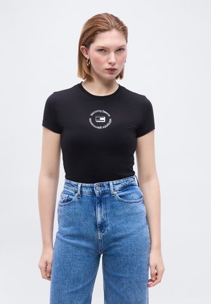 Νεαρή γυναίκα με κοντά κόκκινα μαλλιά που φοράει στενό μαύρο t-shirt με κυκλικό λευκό λογότυπο και ψηλόμεσα μπλε τζιν, στέκεται μπροστά σε απλό φόντο.