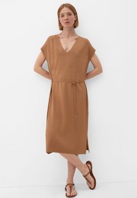 Vrouw in een beige V-hals midi-jurk met een dunne riem, staand op blote voeten in bruine sandalen tegen een witte muur met de armen achter de rug.