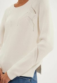 Pull en tricot blanc avec un design texturé et des accents en dentelle, encolure ronde et fentes sur les côtés à l'ourlet.