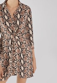 Abito camicia con motivo pitonato nei toni beige, nero e marrone. Presenta un colletto, bottoni e una cintura in vita. Maniche lunghe.