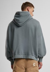 Hooded sweatshirt in soft gray fabric with a loose fit, dropped shoulders, and ribbed cuffs. Features decorative stitching at the hem.