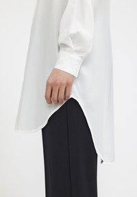 Camicia bianca a maniche lunghe con orlo curvo e polsini con bottoni, abbinata a pantaloni neri. Il tessuto sembra leggero e liscio.