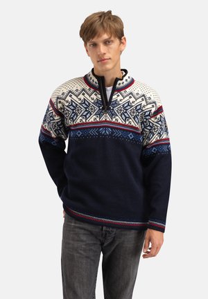 Ung mann iført marineblå og hvit halvglidelåssweater med nordisk mønster og røde detaljer, kombinert med mørkegrå jeans, stående foran hvit bakgrunn.