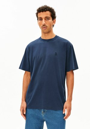Mann mit kurzen lockigen Haaren und Schnurrbart, der ein schlichtes dunkelblaues T-Shirt und blaue Jeans trägt, steht vor einem weißen Hintergrund.