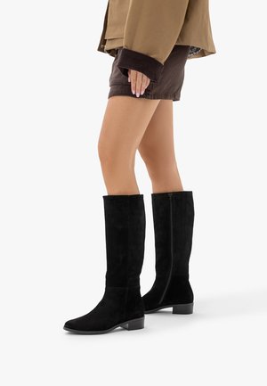 Jambes portant des bottes noires montantes, un short marron foncé et une veste beige avec des poignets foncés, vues de profil sur un fond blanc.