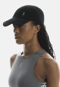 Sort sports cap med perforeringer, med et reflekterende logo. Båret af en person i en grå atletisk top, foran en ensfarvet baggrund.