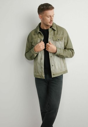 Mężczyzna dopasowujący jasnooliwkową kurtkę dżinsową na czarnej koszuli, ubrany w ciemne dżinsy, stojący na tle jednolitego, jasnego tła.