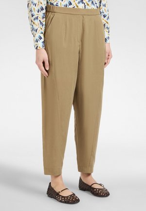Femme portant un pantalon beige fuselé, une chemise à manches longues à motifs et des ballerines noires tissées, debout sur un fond blanc uni.