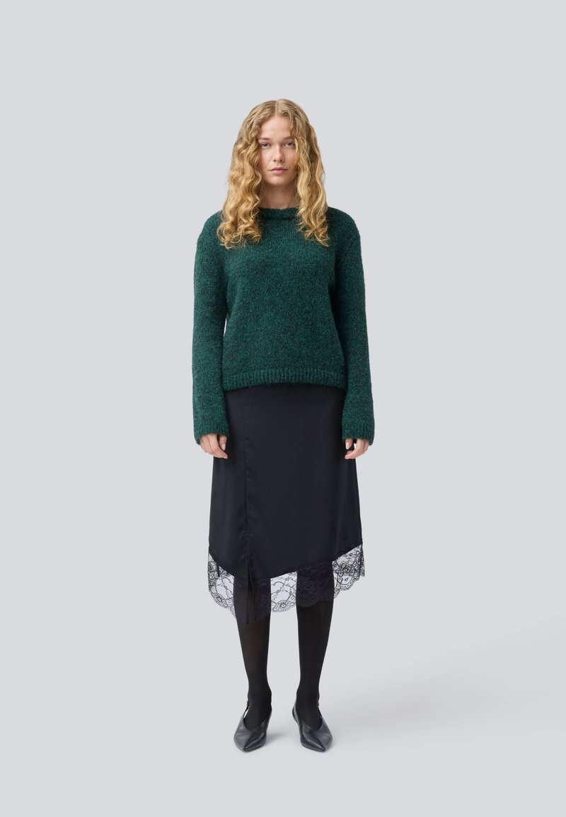 Jeune femme avec de longs cheveux blonds bouclés portant un pull vert foncé, une jupe noire avec un ourlet en dentelle, des collants noirs et des chaussures à talons noires.