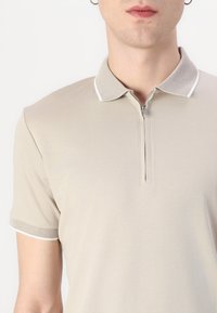 Beige polo skjorte med glidelås ved halsen, teksturert krage og hvite detaljer på kragen og ermene. Glatt stoff, tettsittende design.